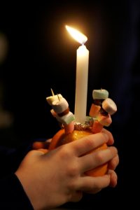 Photograph of a child's hands holding a Christingle - an orange with a candle and other items stuck in it
