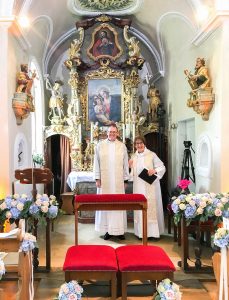 A photograph of a man and a woman, dressed in white albs in a highly decorated chapel